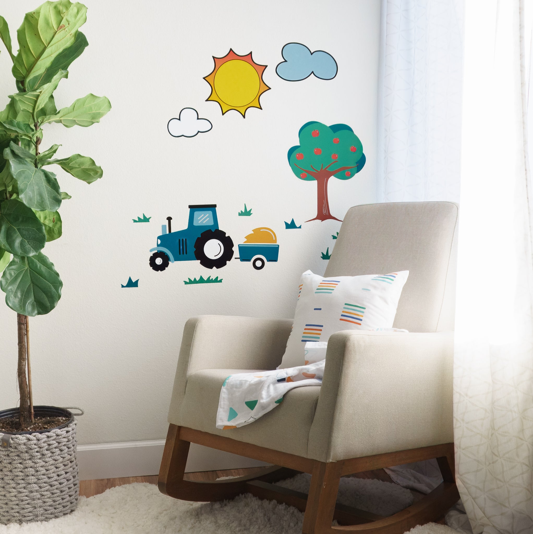 Baby nursery with plant on left, rocking chair and colorful throw pillow and blanket on right in front of a window with white fluffy rug. On the white wall behind, there is a farm scene of wall decals including an apple tree, grass, tractor with hay and wagon, clouds and sun.
