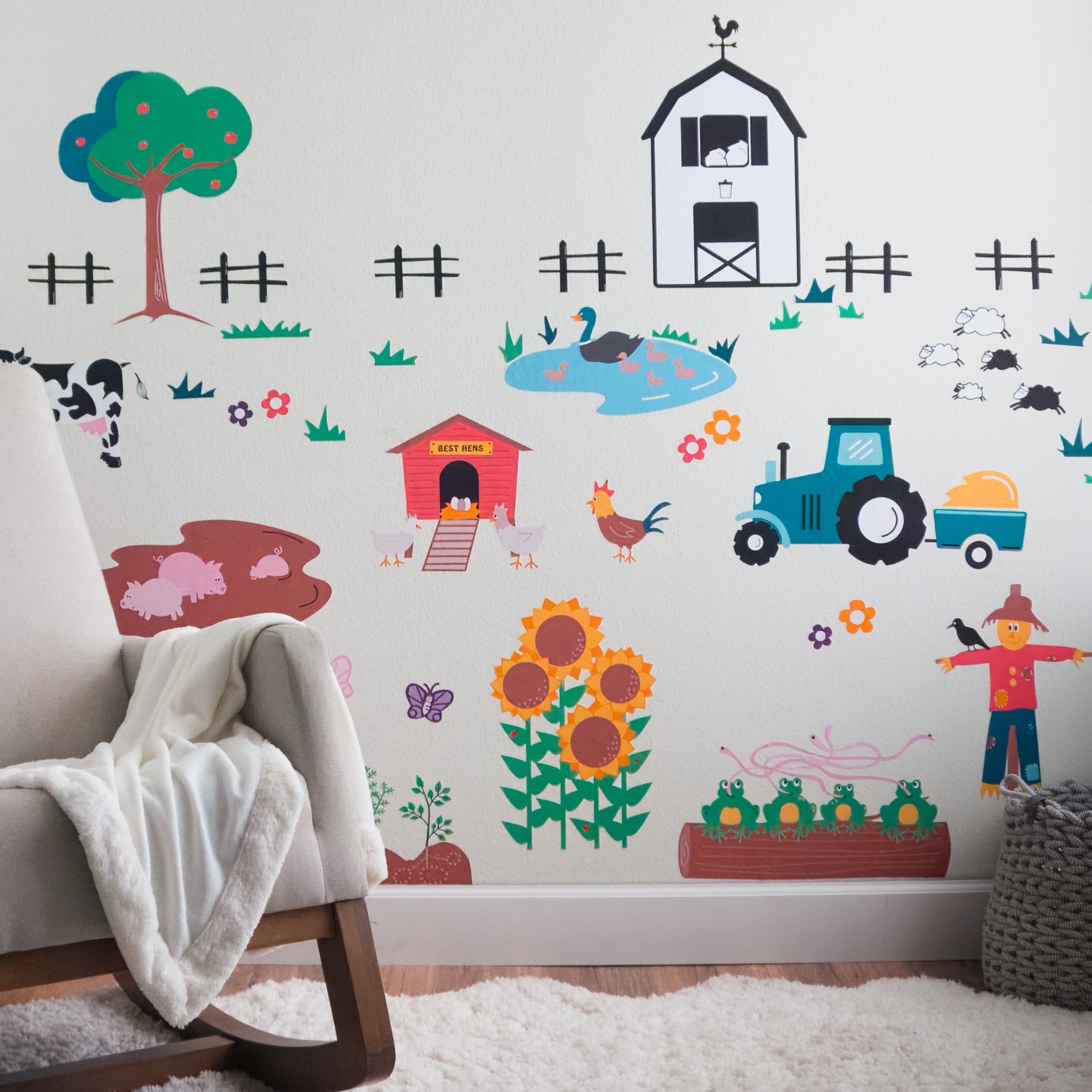 A kid's room wall featuring a farm mural made of wall decals including a hen house with hens and eggs, pigs in a mud pit, a cow, sunflowers, a garden, frogs on a log, scarecrow, tractor, sheep, a duck in a pond, an apple tree, fences, grass, flowers and butterflies. In front is a rocking chair with a white blanket draped on the side, a fluffy white rug and a grey storage basket.