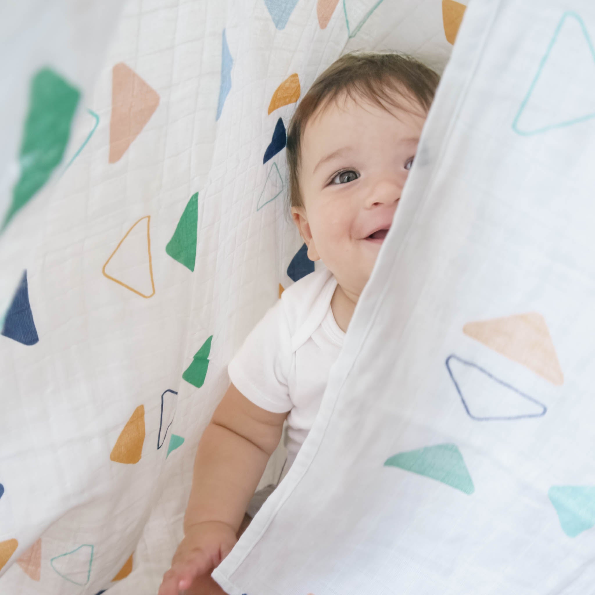 A baby is half covered by a white muslin blanket with colorful triangles all over. He is smiling.