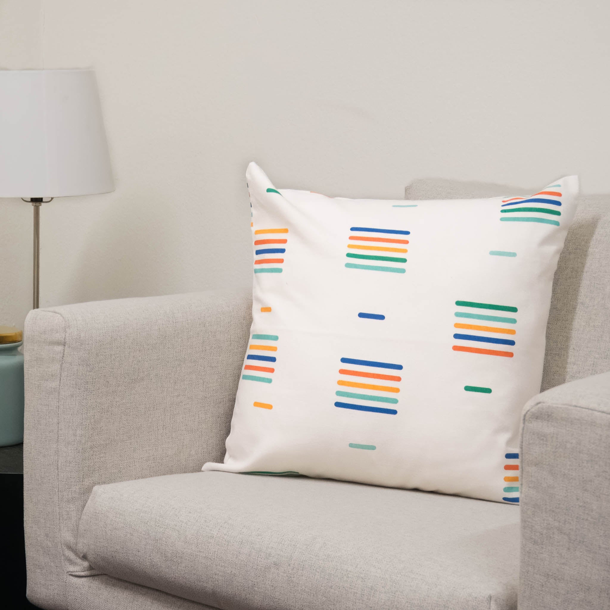 A chair with a throw pillow on top. The throw pillow is white with a linear repeated pattern in different bright colors.