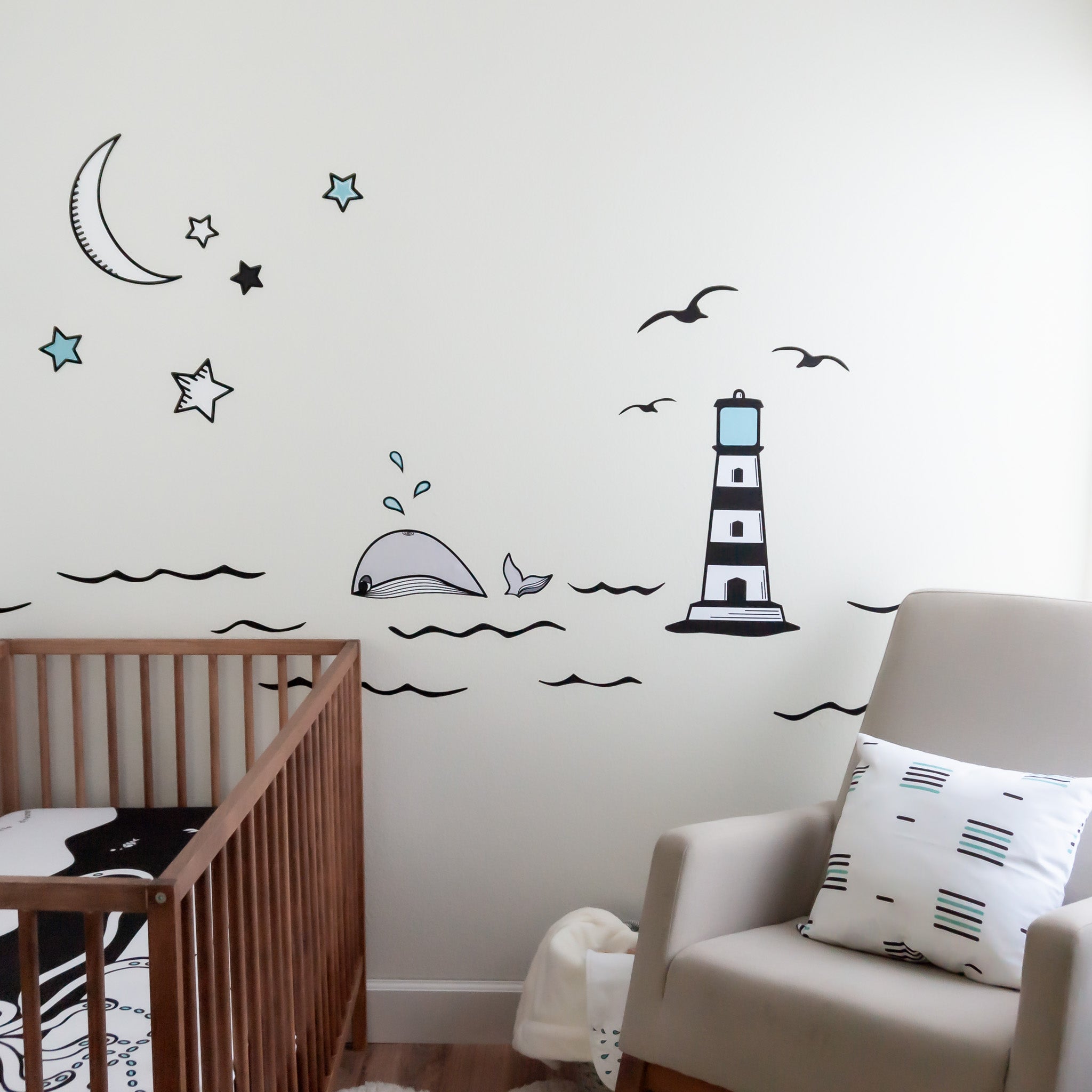 A nursery with a rocking chair on the right, wooden crib on the left with a black and white crib sheet. The wall has an ocean mural with a lighthouse, whale, moon and stars.