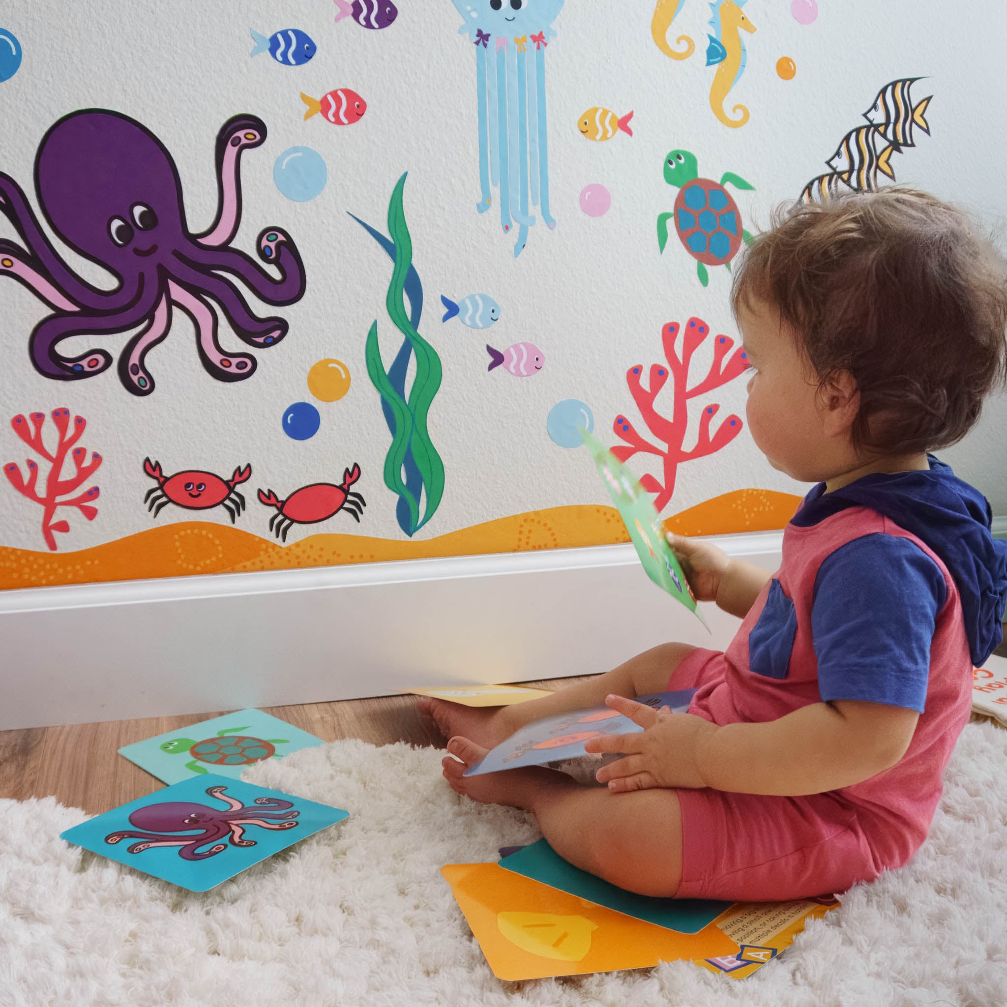 A toddler sits on the right holding colorful activity cards that match the ocean mural on the wall in front of him.