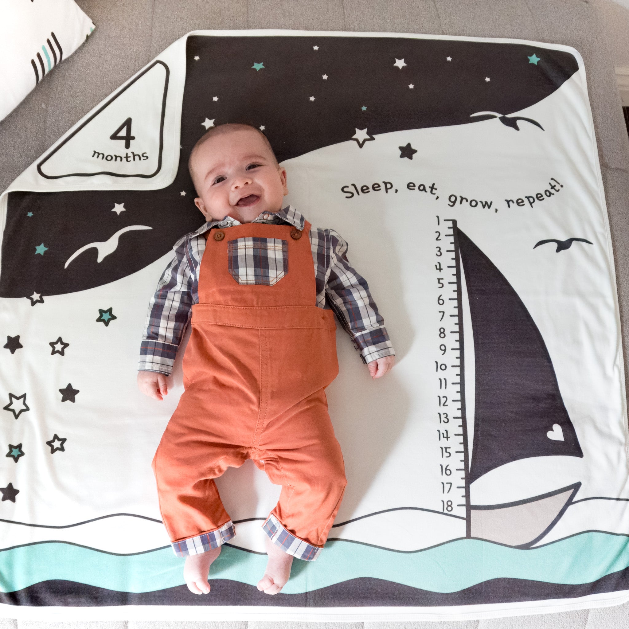 A baby wearing overalls lying on a black and white blanket with an ocean scene, smiling.