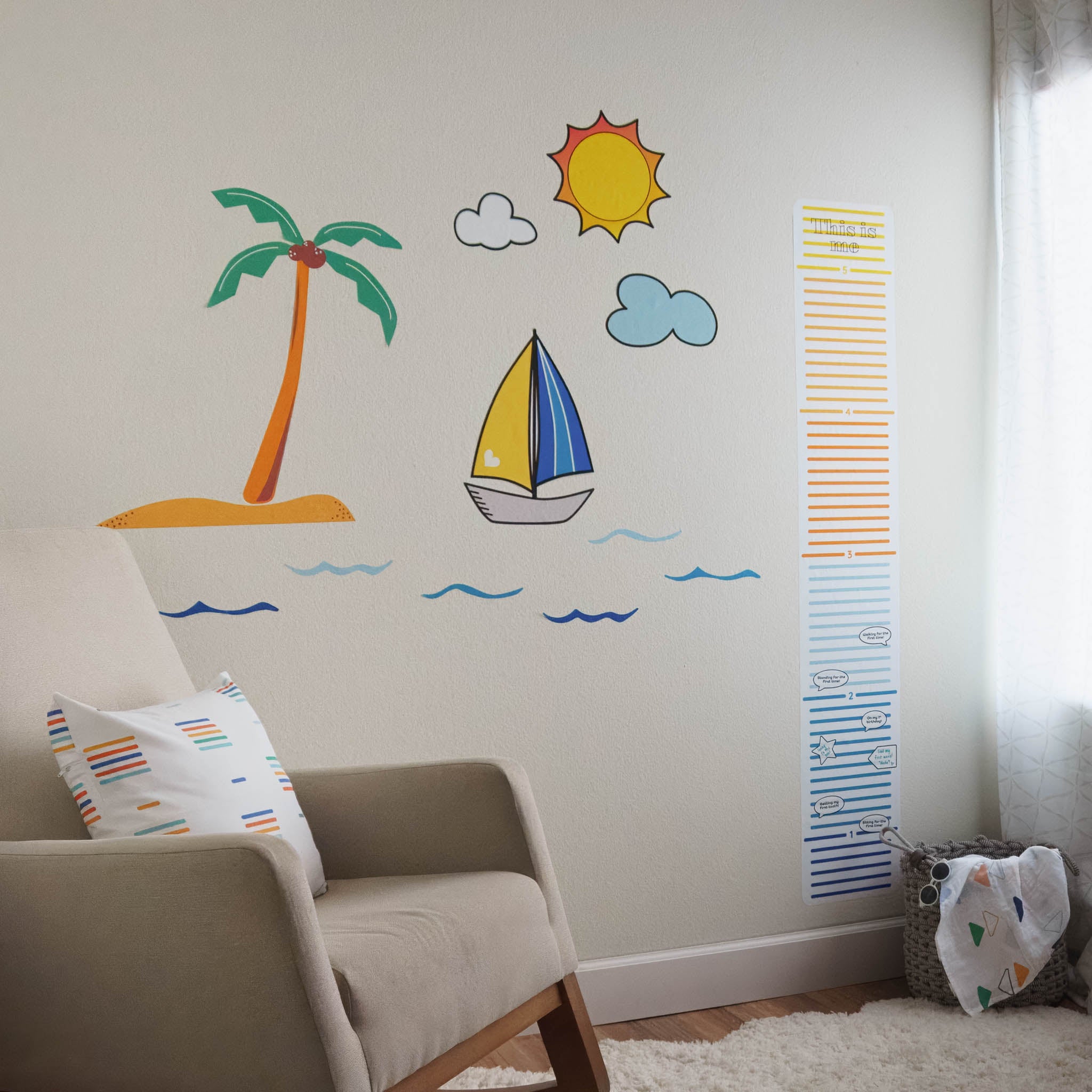 A baby's nursery with rocking chair on the left side, with a multi-colored throw pillow on it. There is a scene on the wall in wall stickers, with blue waves, a tropical island, sailboat, sun and clouds. There is a colorful growth chart on the white wall as well. On the ground there is a white rug and a basket with a blanket.