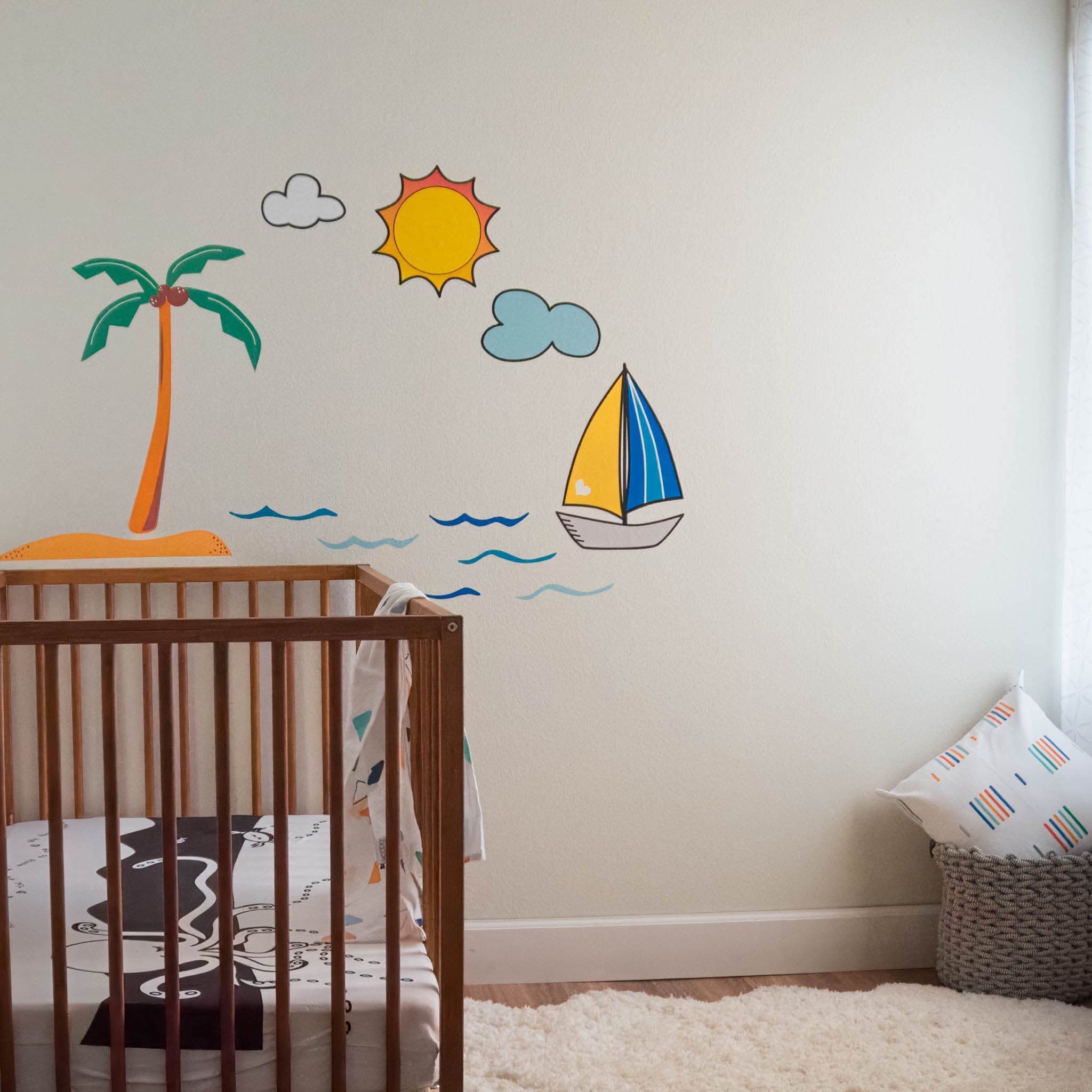 A nursery room with a crib on the left, a window on the right and a wall mural of a tropical ocean scene including a palm tree, sailboat and sun with clouds.