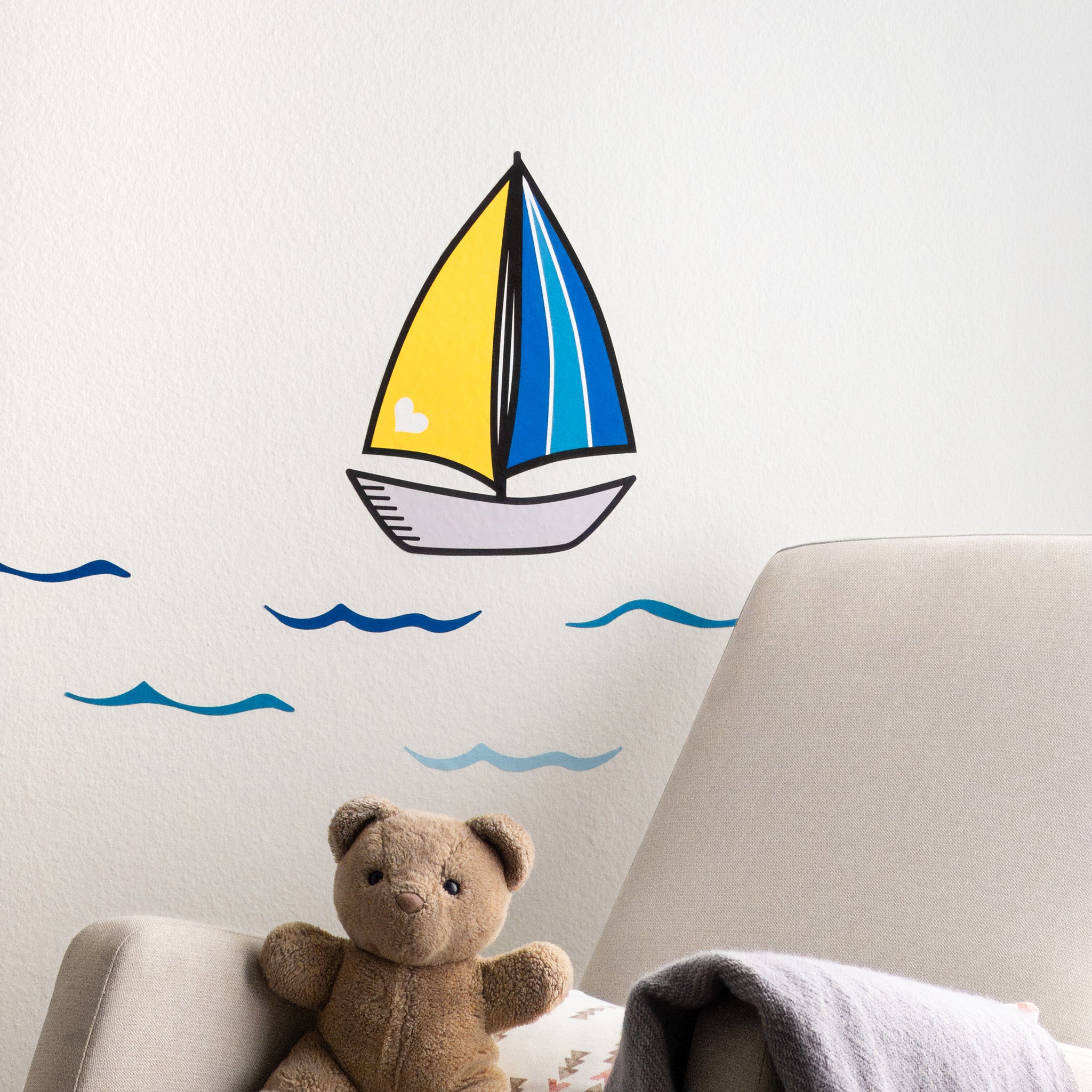 A white wall with a sailboat wall decal that has a yellow sail on one side and striped blue sail on the other. The body of the boat is grey. There are bright blue waves underneath. On the right side, there is a partial view of a rocking chair with blanket and teddy bear.