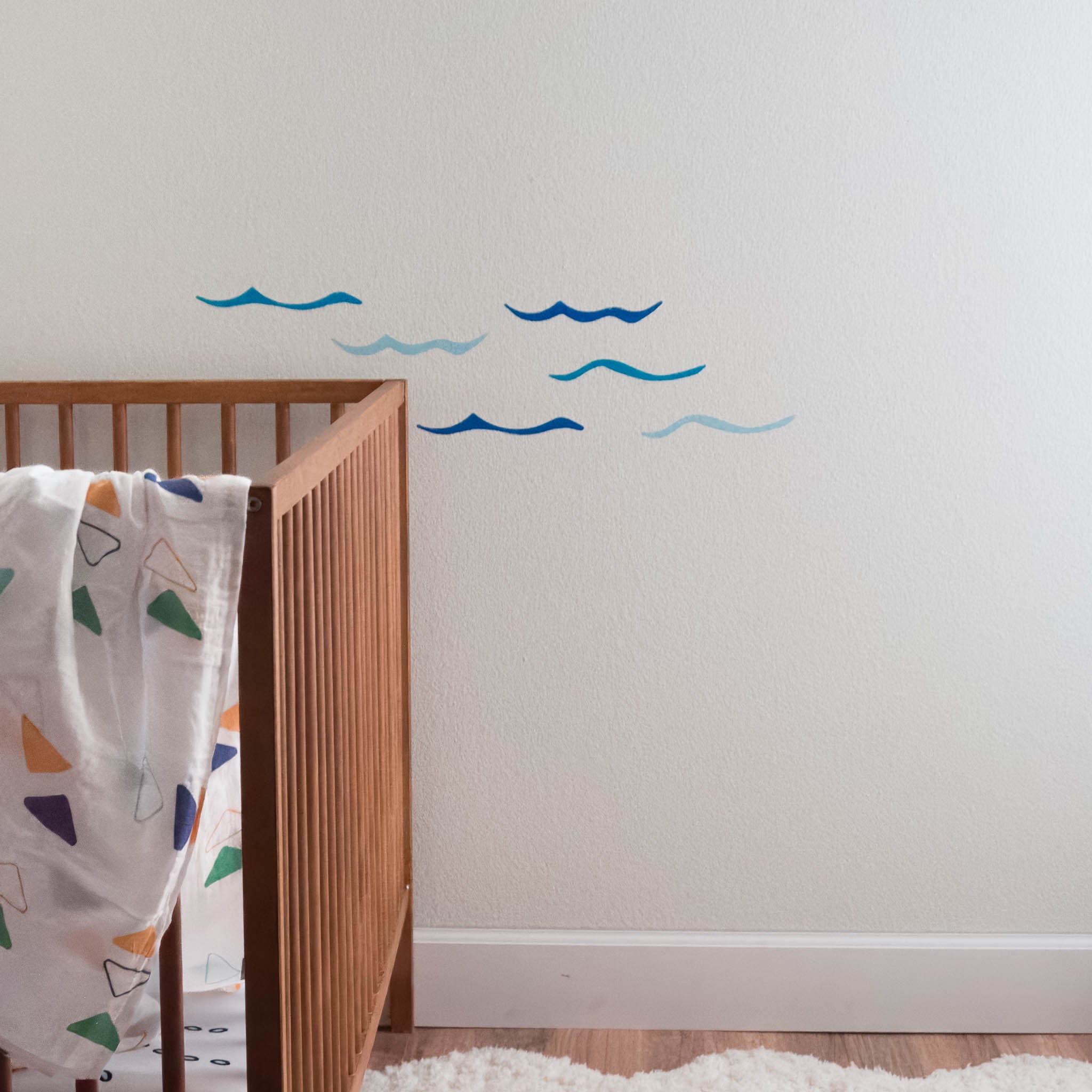 A white wall with a wooden crib in front with a muslin blanket draped over one side. On the wall are a set of 6 bright blue ocean wave wall decals in varying shades of blue.