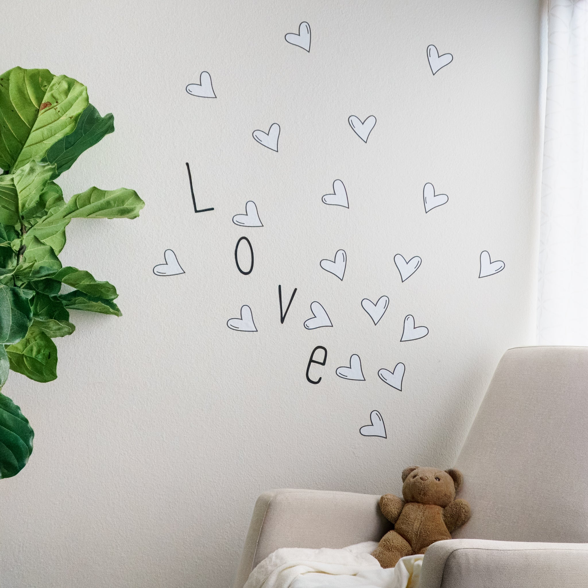 Baby nursery with green plant on left with rocking chair and teddy bear on the right. On the wall behind is the word "love" in a black, modern font surrounded by black and white heart wall decals.
