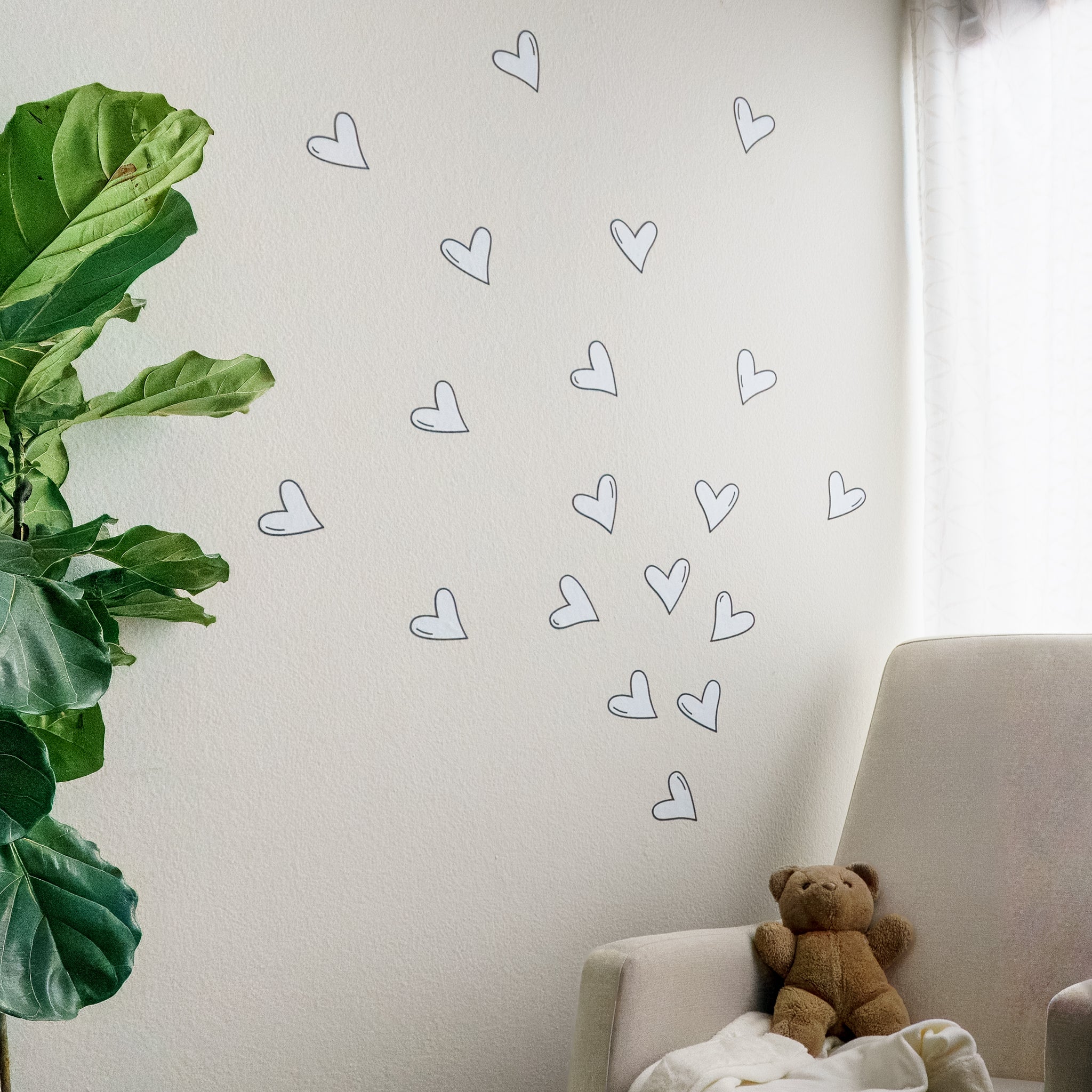 Baby nursery with green plant on left, rocking chair with teddy bear on right. A white wall behind the rocking chair has multiple white heart wall decals with black outlines, arranged as though coming from the chair.
