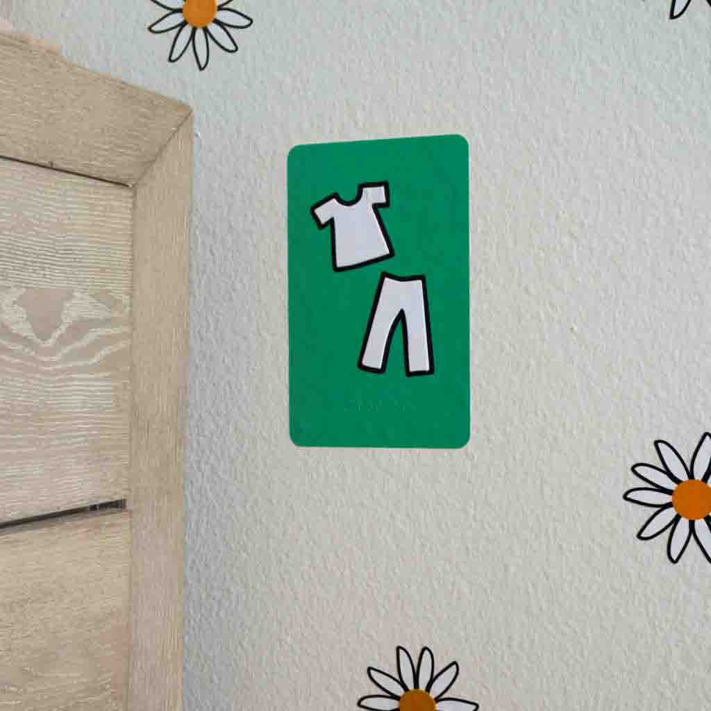 A green wall decal with a pants and shirt in white and braille applied on the wall next to a dresser