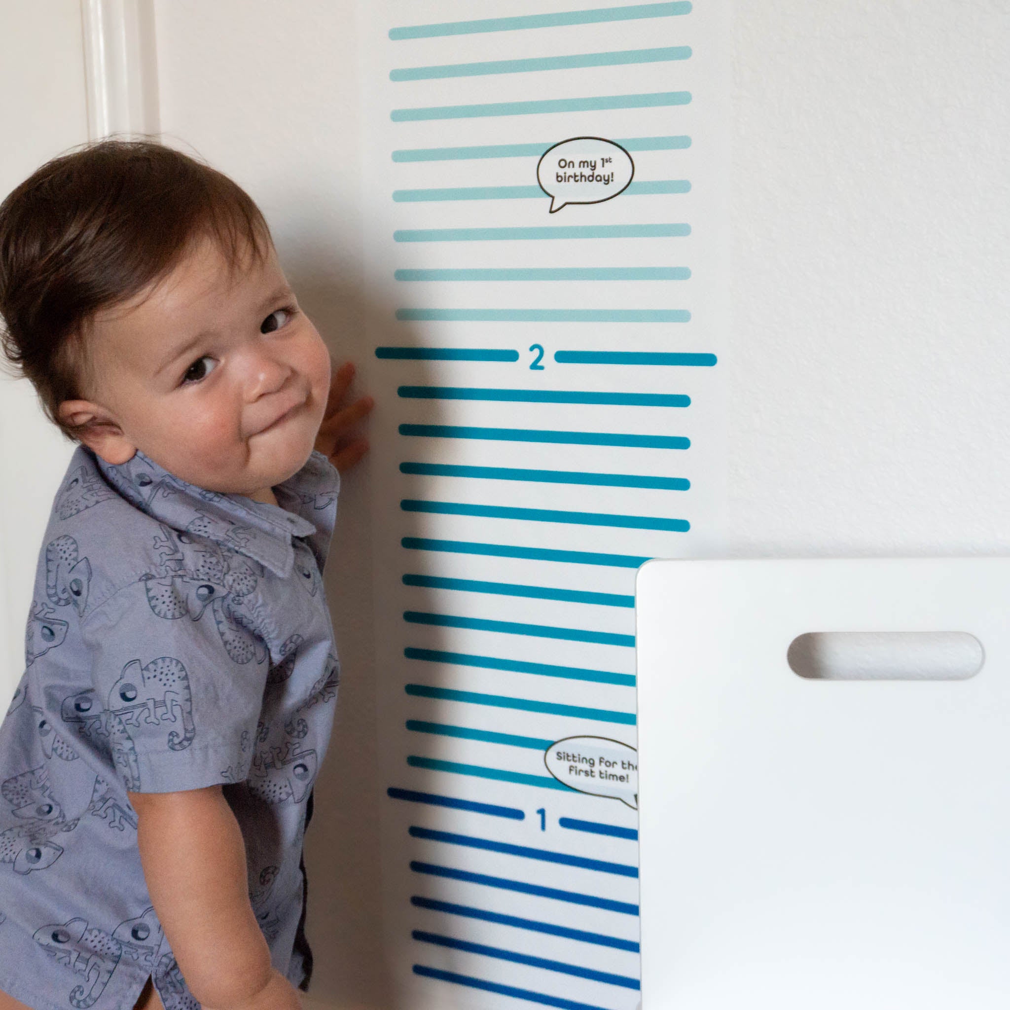 A growth chart wall decal with multicolored stripes on the wall with a toddler standing next to it wearing a blue shirt.