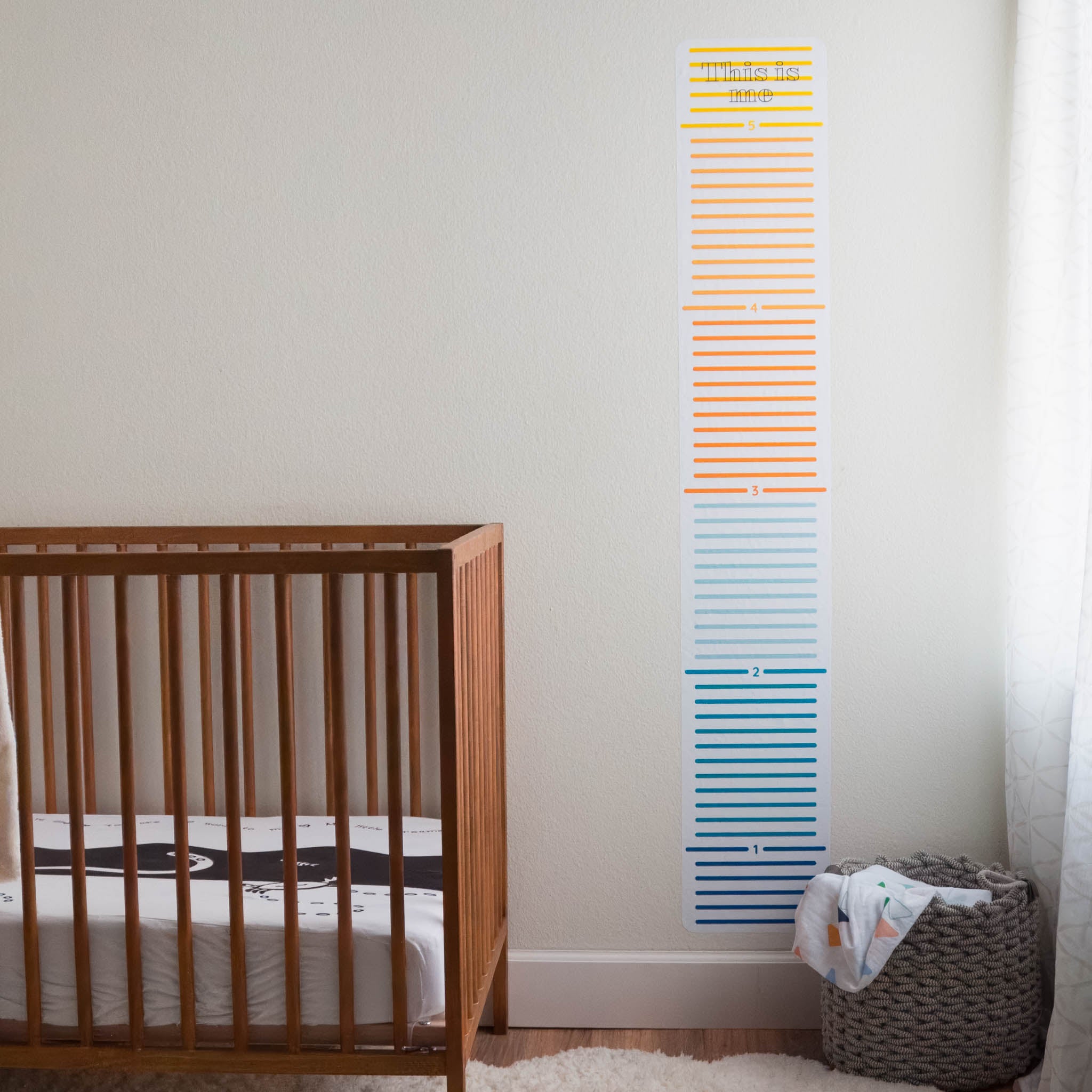 A wooden crib is on the left, to the right is a window. On the white wall in between there is a colorful growth chart wall decal for a child. The top says "This is me".
