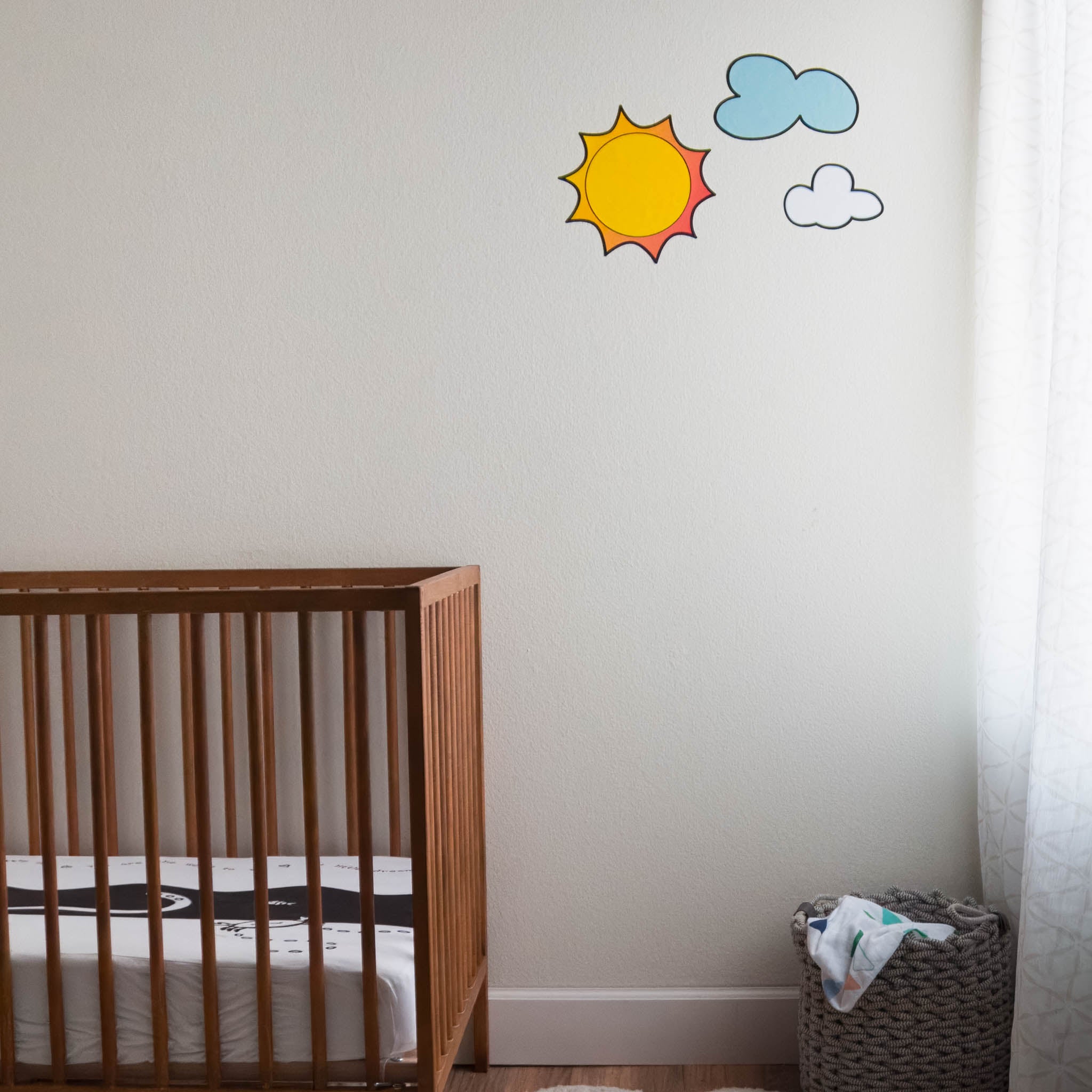 A nursery room with wooden crib on the left and window on the right. On the wall there is a sun and clouds in wall stickers.