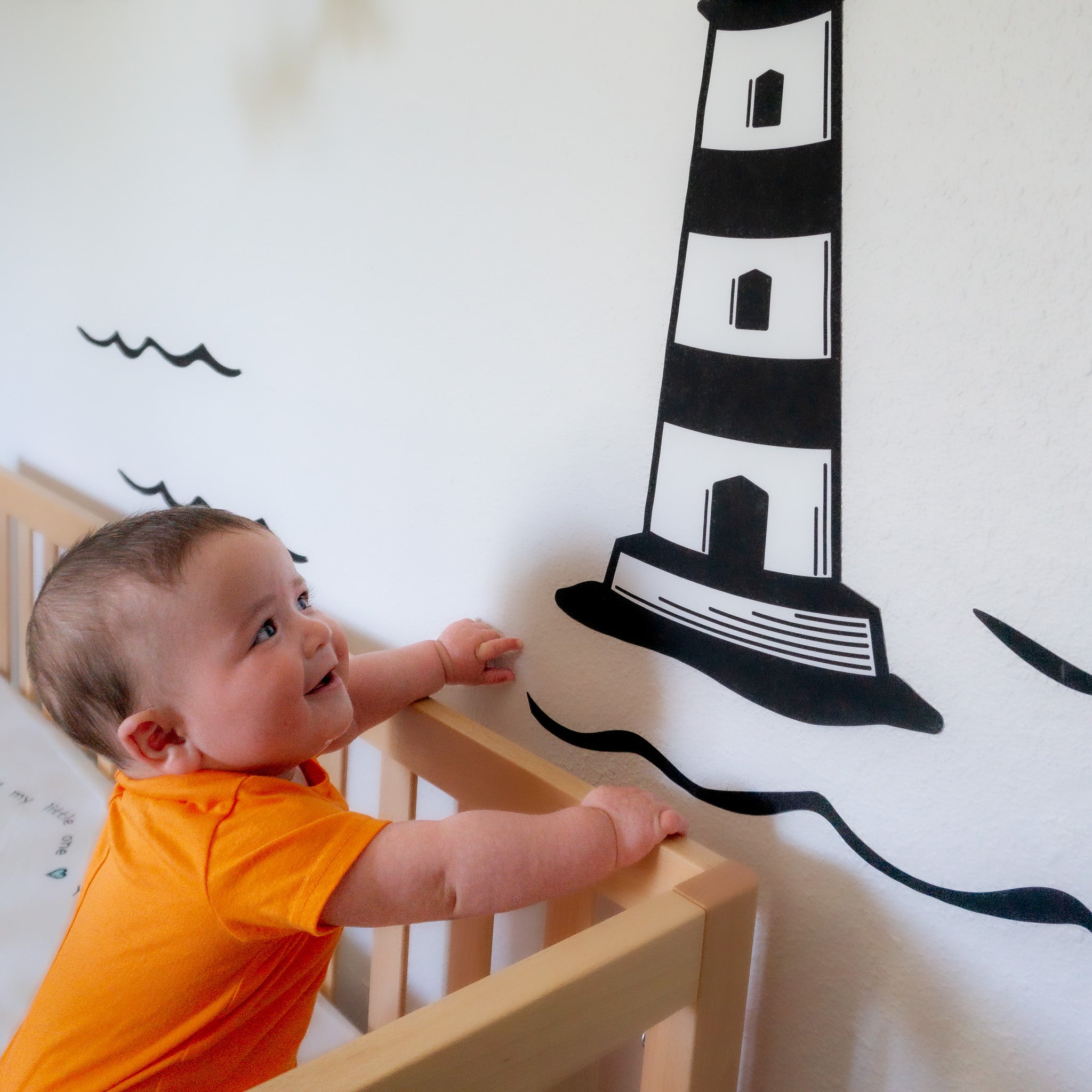 A baby wearing an orange onesie on the left looking and smiling at a wall sticker of a black and white lighthouse.