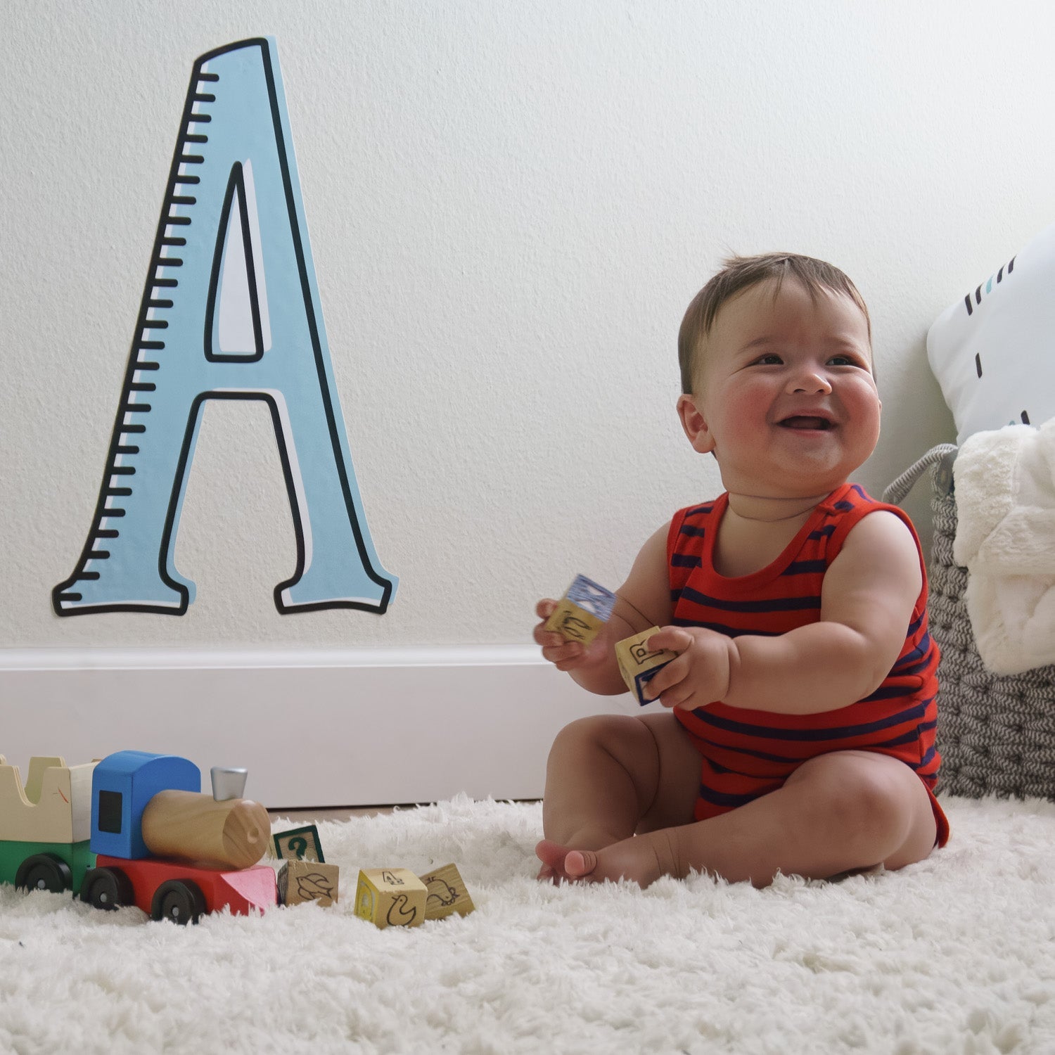 A scene with a baby boy laughing and playing with blocks on a white rug with a large letter "A" wall decal in blue and black in the background.