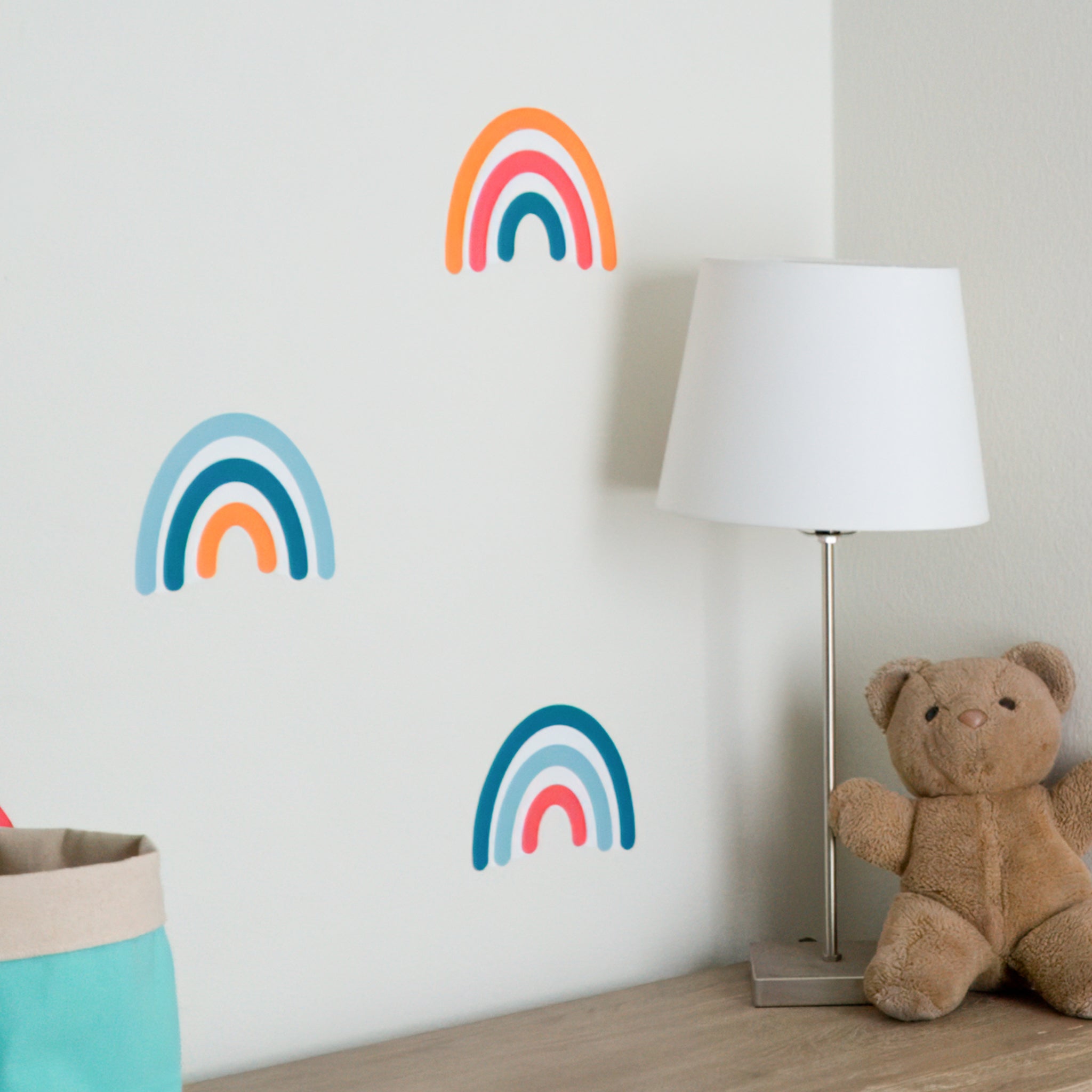 Rainbow wall decals on wall with a teddy bear, lamp and cloth storage basket on a dresser in front