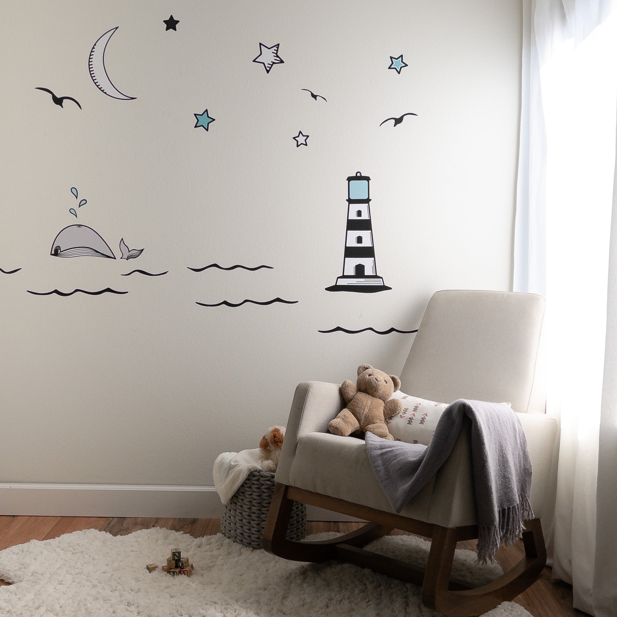 Baby's nursery with rocking chair, teddy bear and white rug. On the wall is an ocean mural with a lighthouse, whale, black waves and moon and stars.