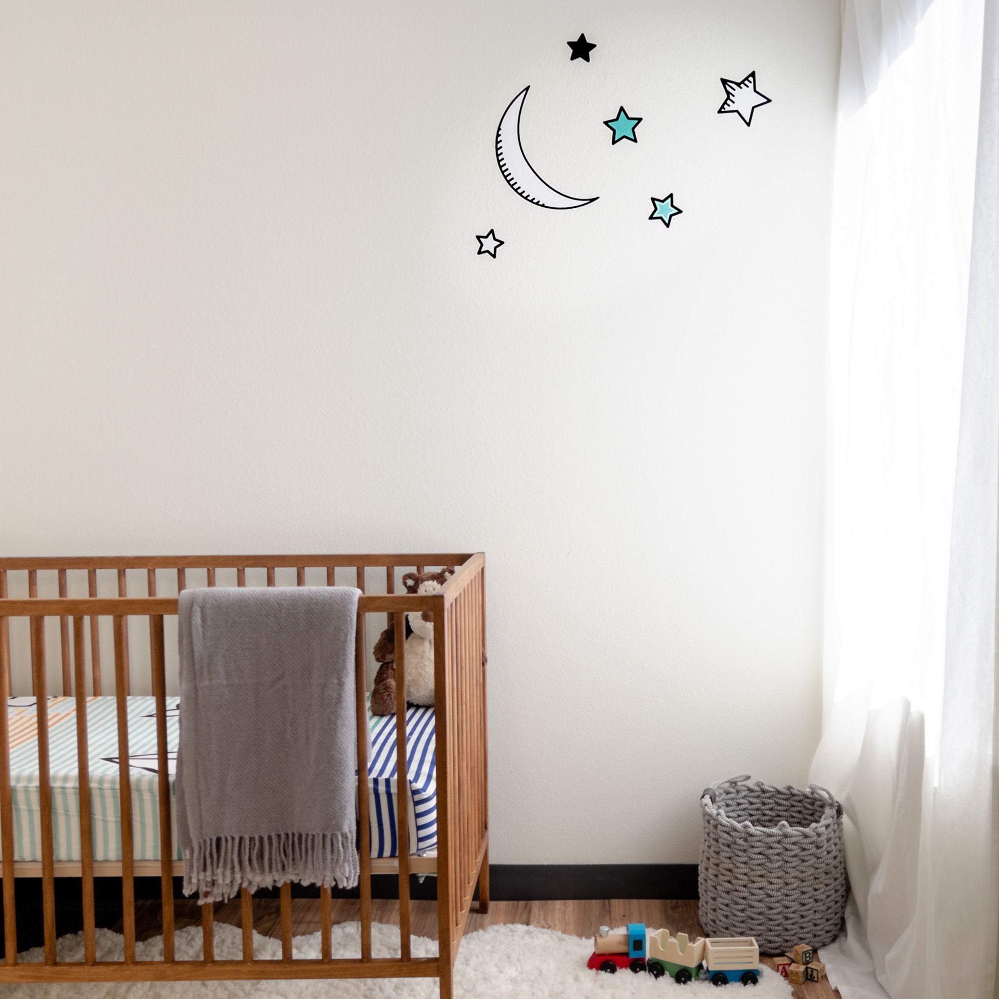 A baby's nursery with a wooden crib on the left, a window on the right and moon and star wall decals on the wall.